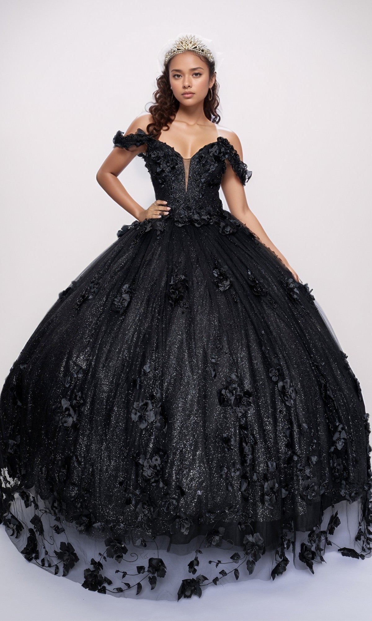 A woman wears the Quinceanera Dress Q2444 By Nina Canacci in black, featuring off-shoulder 3D floral appliques and a glitter tulle skirt, paired with a tiara, as she poses against a plain light background.